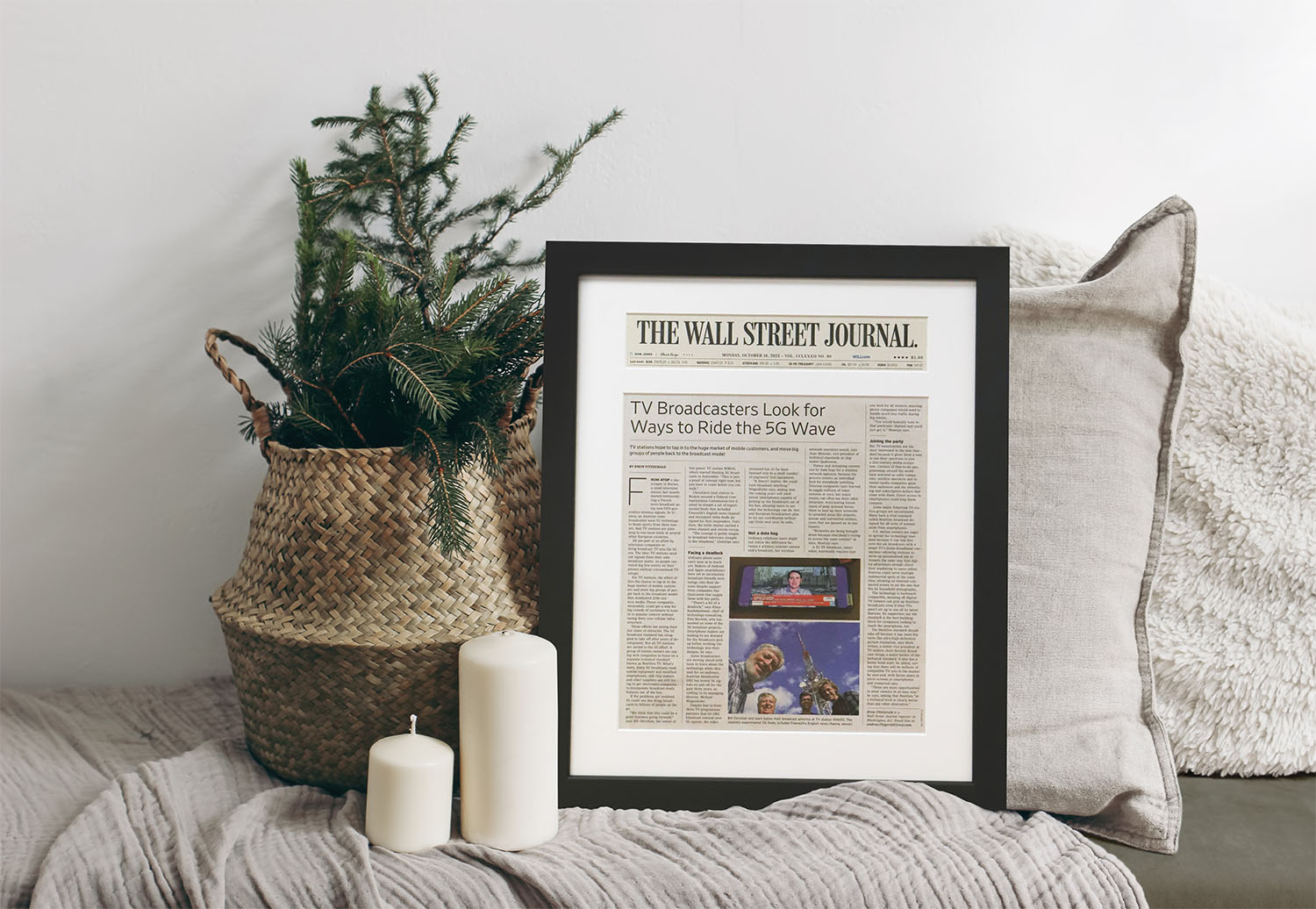 Framed Wall Street Journal newspaper article displayed in a modern black frame with white matting, resting on a cozy sofa beside candles and holiday greenery.