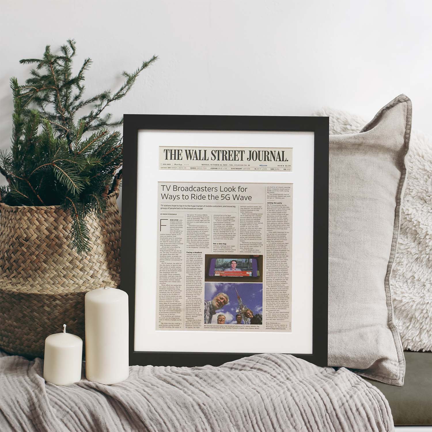 Framed Wall Street Journal newspaper article displayed in a modern black frame with white matting, resting on a cozy sofa beside candles and holiday greenery.