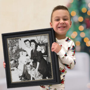 Smiling boy in holiday pajamas holding a framed vintage black-and-white family Christmas photograph in front of a glowing tree.